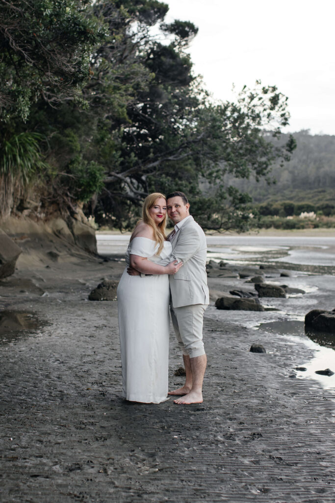 Romantic couple holding hands on rocky shoreline at Kakamatua Inlet, West Auckland engagement photography.