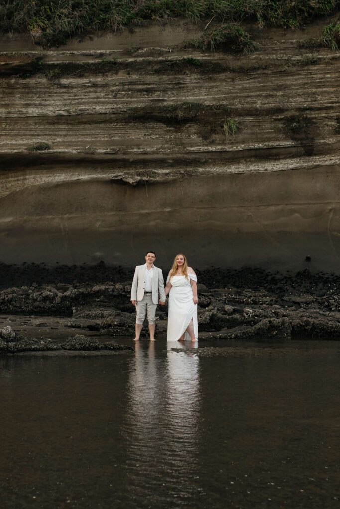 Romantic couple holding hands on rocky shoreline at Kakamatua Inlet, West Auckland engagement photography.