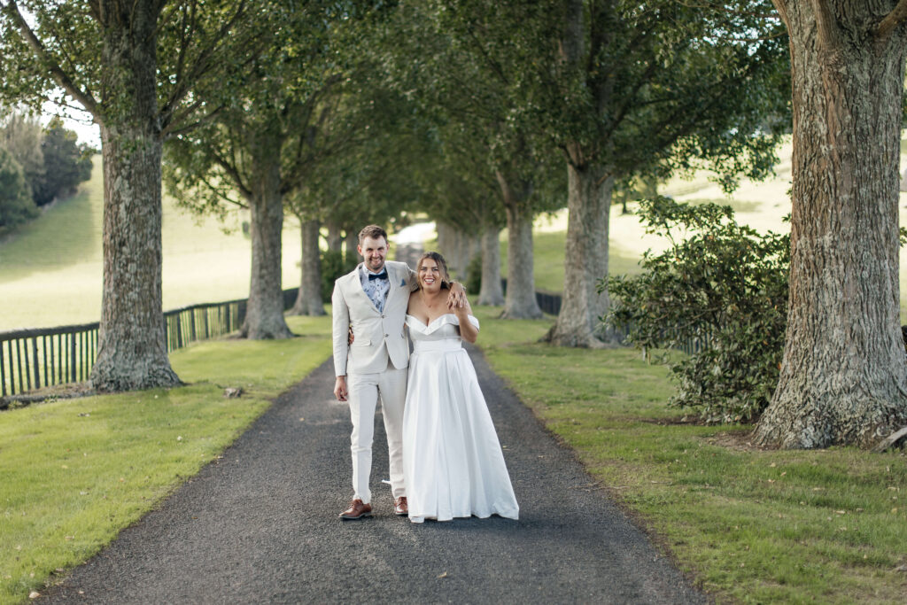 Bride and groom walking hand in hand down the tree-lined driveway at The Red Barn Karapiro