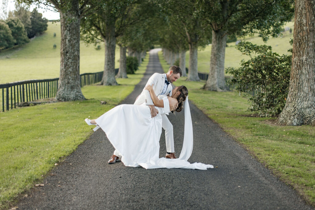 Bride and groom walking hand in hand down the tree-lined driveway at The Red Barn Karapiro