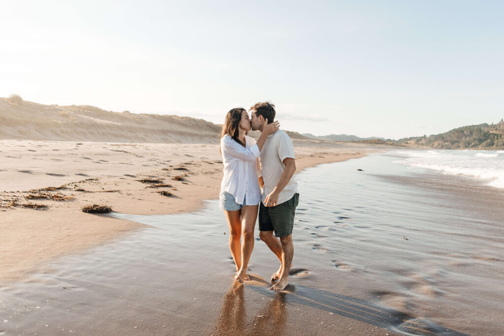 Candid moment of couple embracing at sunset with waves in the background – Coromandel engagement photographer