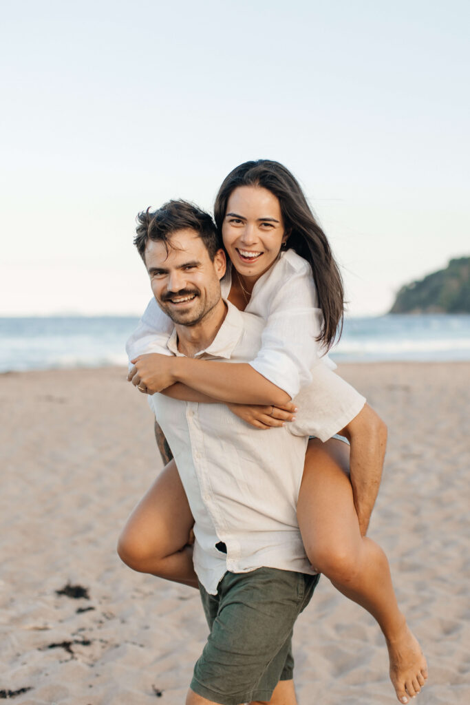 Candid moment of couple embracing at sunset with waves in the background – Coromandel engagement photographer