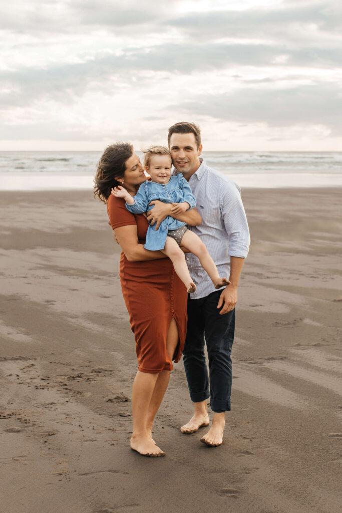 Parents holding daughter on windy Bethells Beach evening