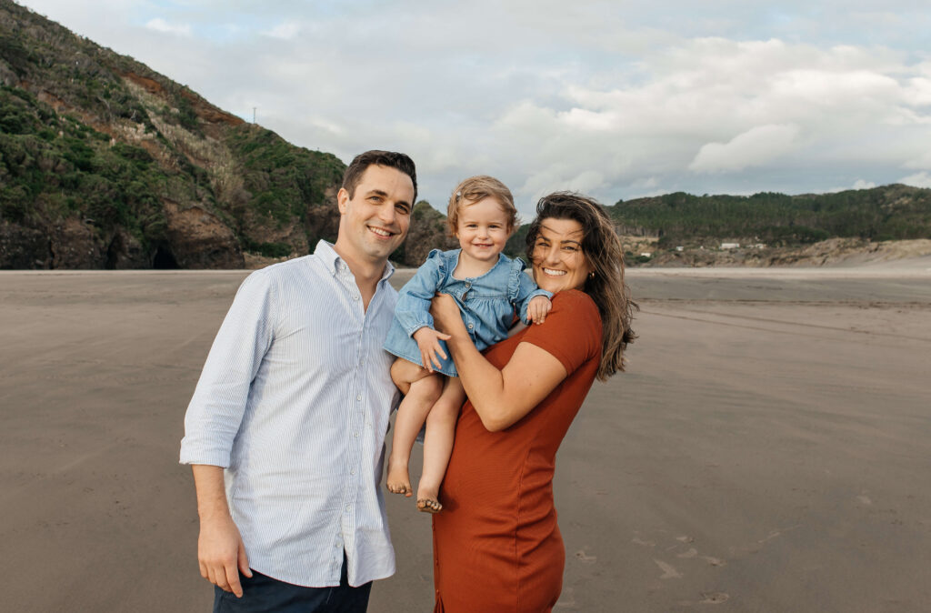 Parents holding daughter on windy Bethells Beach evening